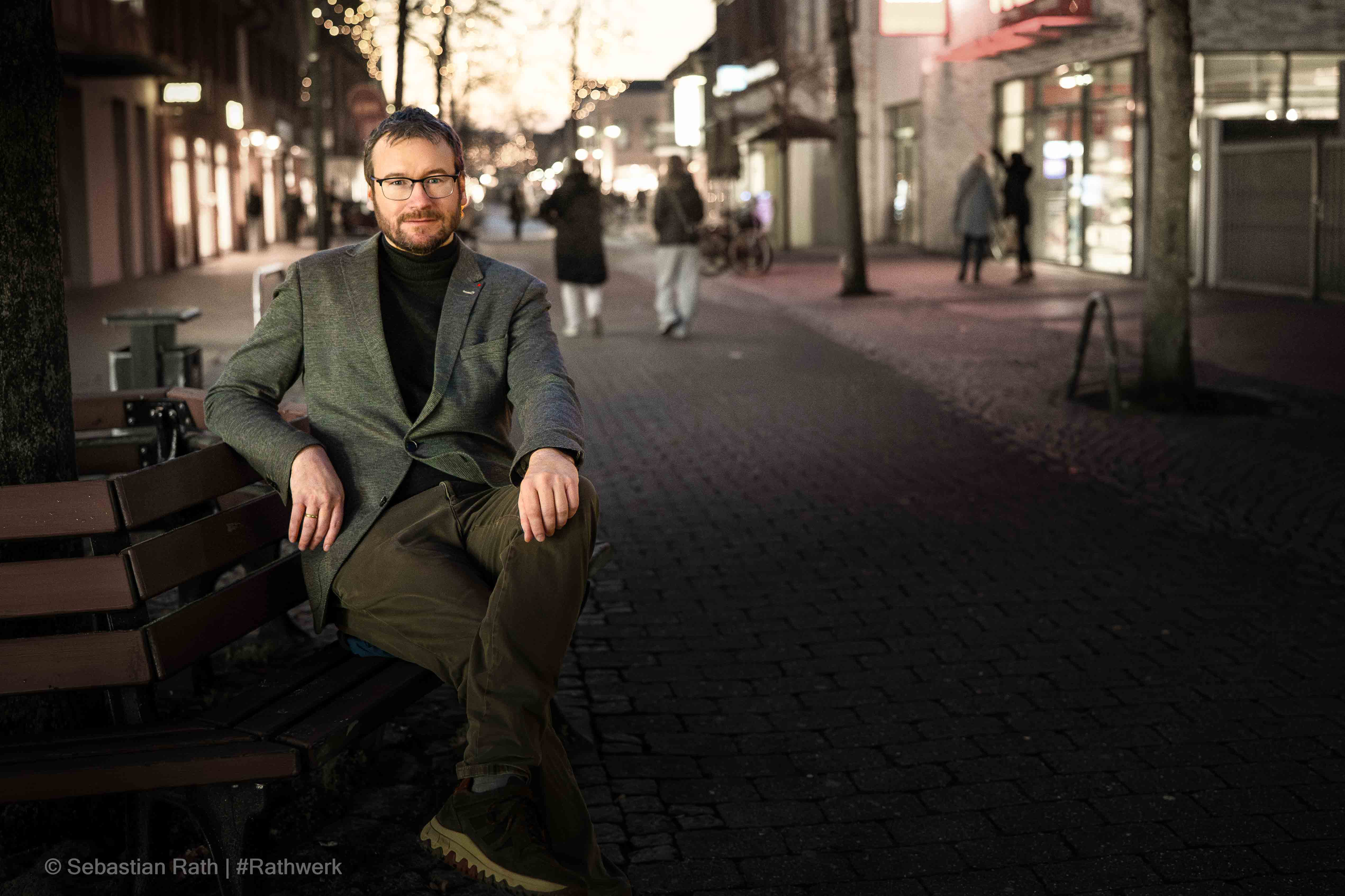 Sebastian Rath sitzt auf einer Holzbank in einer Fußgängerzone während der Abenddämmerung. Er trägt ein graues Sakko über einem dunklen Rollkragenpullover, eine Brille und blickt direkt in die Kamera. Der Hintergrund zeigt eine belebte Einkaufsstraße mit warmen Lichtern.